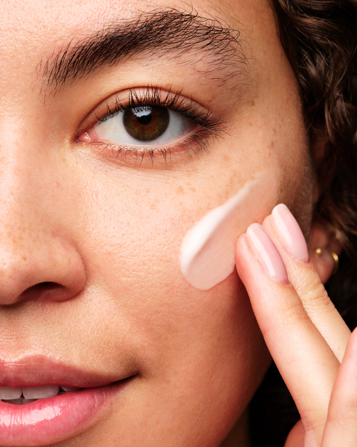 Close-up of a woman's face with a dollop of light pink cream foundation being blended onto her cheek by her fingers.