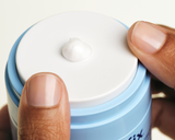 Hands pumping the Skinfix Ceramide Prime Whipped Cream jar, with a small dollop of white cream resting on the lid.