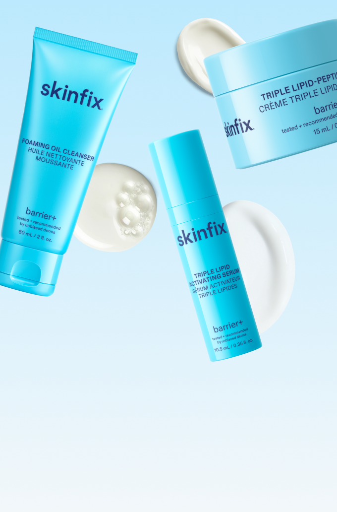 Three skincare products from skinfix barrier+ arranged on a light blue background: a foam cleansing tube, a serum bottle, and a cream jar, with white cream swirls and pearl-like droplets as styling props.