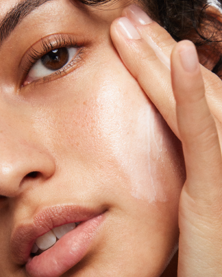 Close-up of a person applying moisturizer to the cheek looking into the camera.