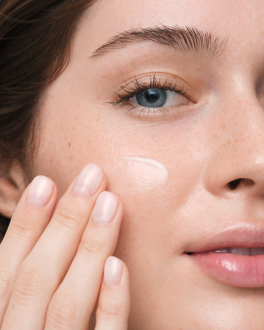 Close-up of a woman's face with a dollop of cream product on her cheek, her hand touching her skin, showing a clean skincare application.