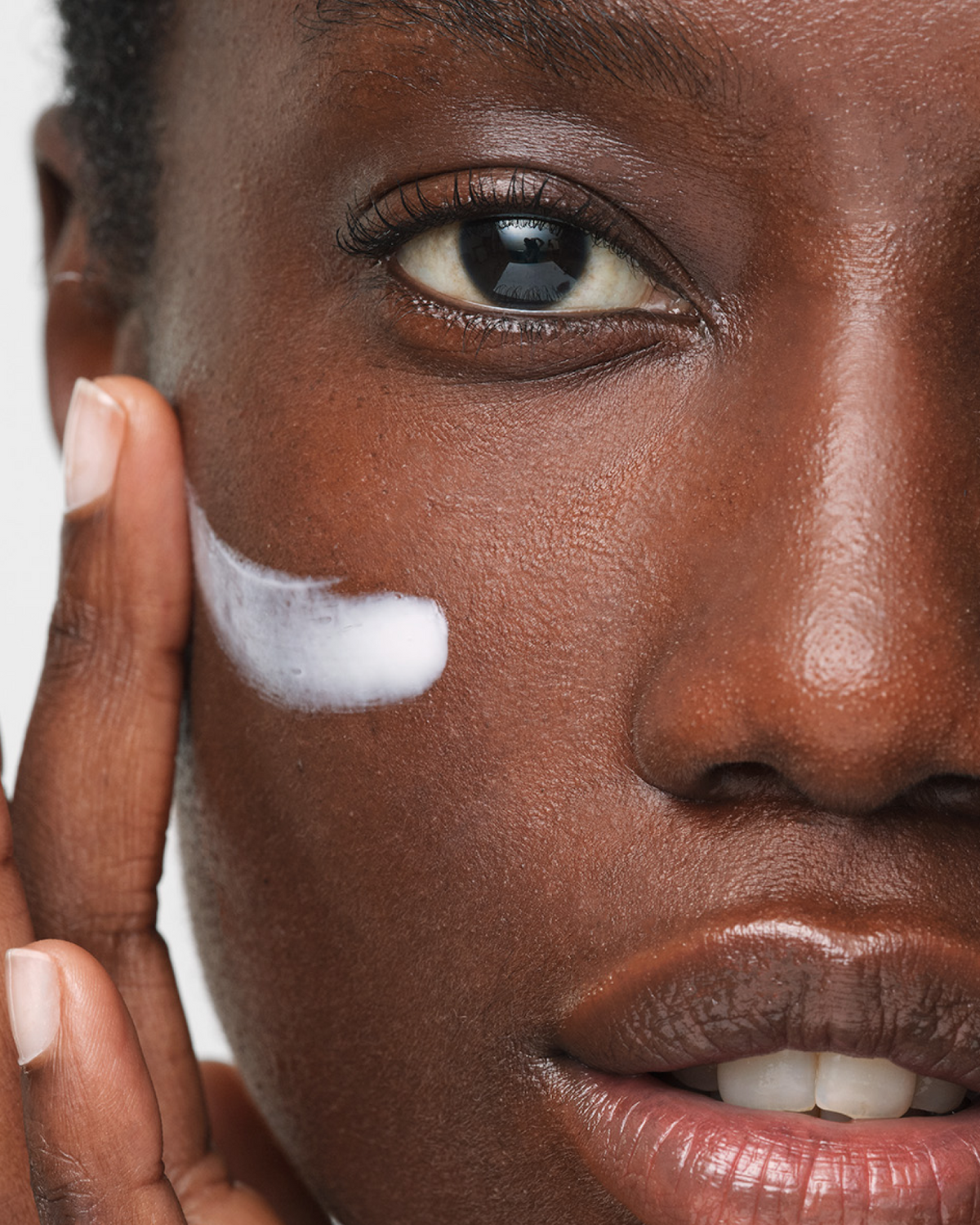 Close-up of a woman's face with white cream applied to her cheek, touching her face with her fingers against a light gray background.