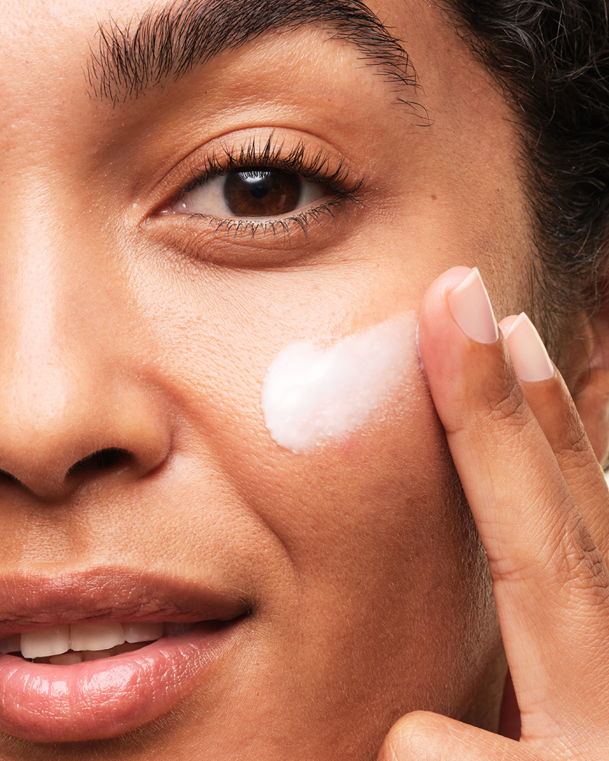 Woman looking into camera applying Skinfix Brighten + Firm Vitamin C Serum to right cheek.